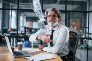 Throwing money. Mature businessman with grey hair and beard in formal clothes is in the office